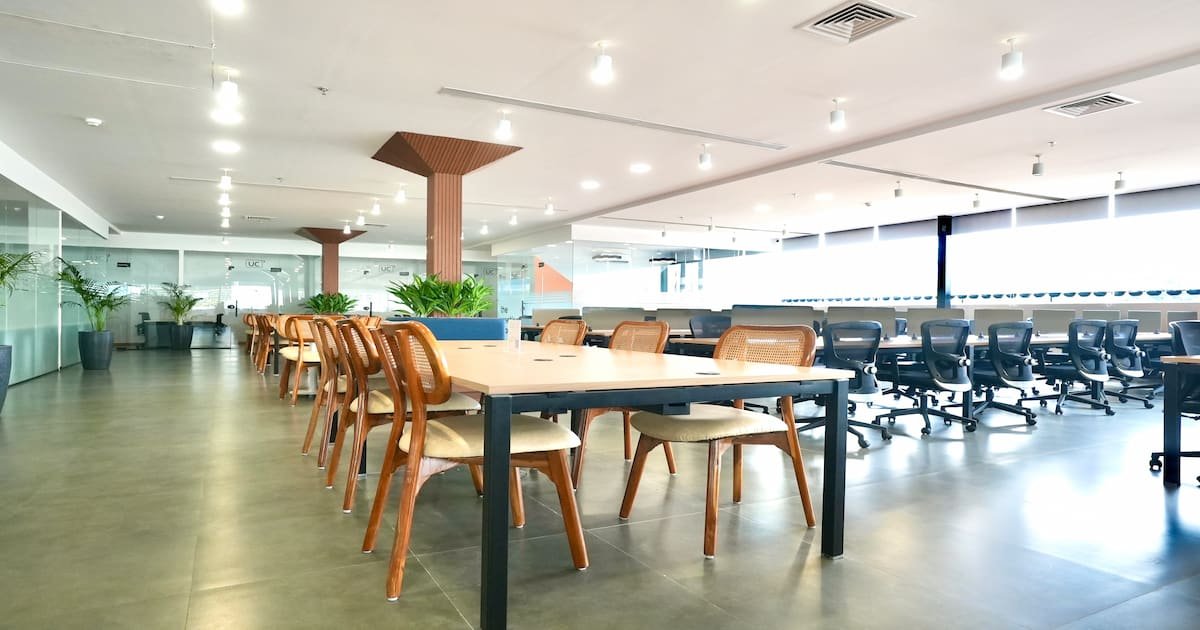 Spacious coworking space with wooden chairs, ergonomic office chairs, large desks, and indoor plants under bright ceiling lights.